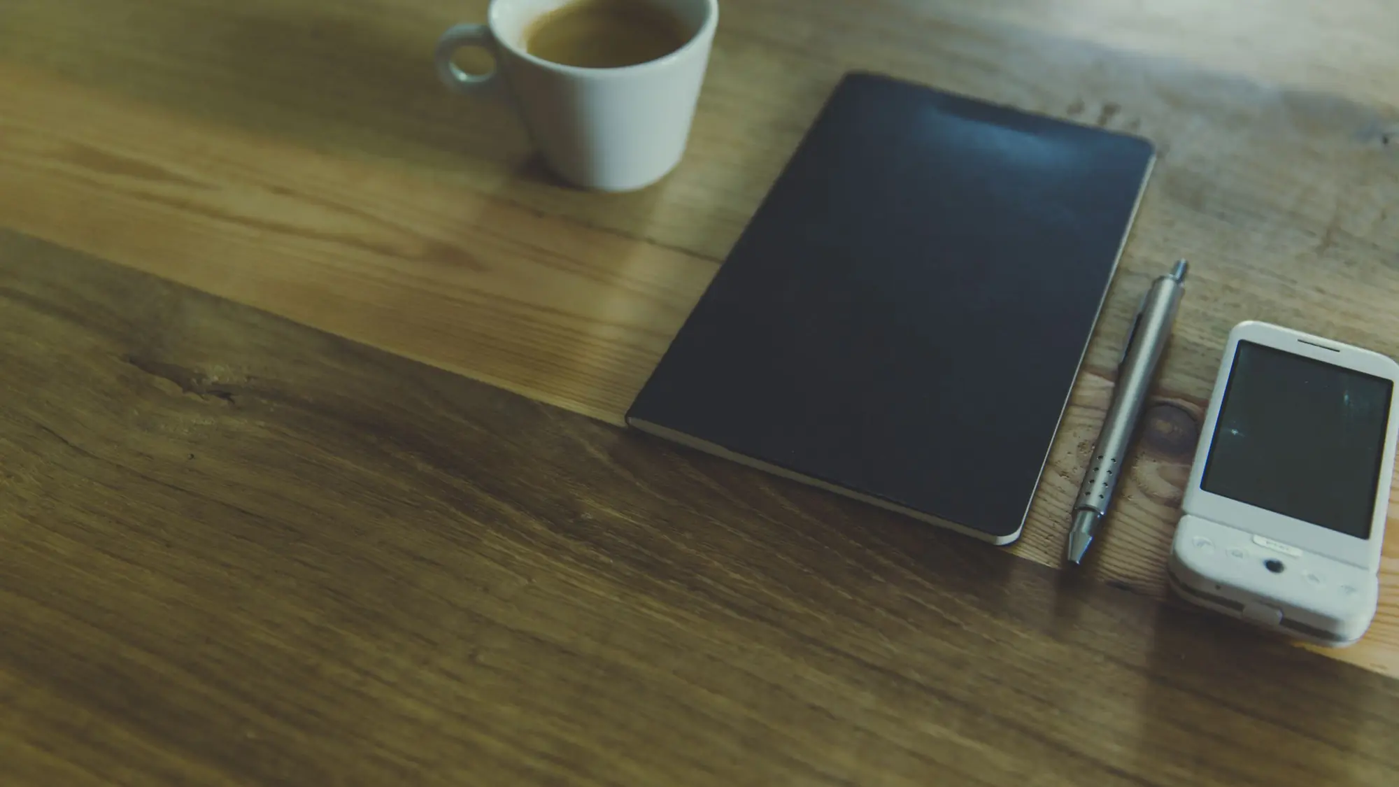 Taza de café, cuaderno cerrado, bolígrafo y teléfono sobre una mesa de madera