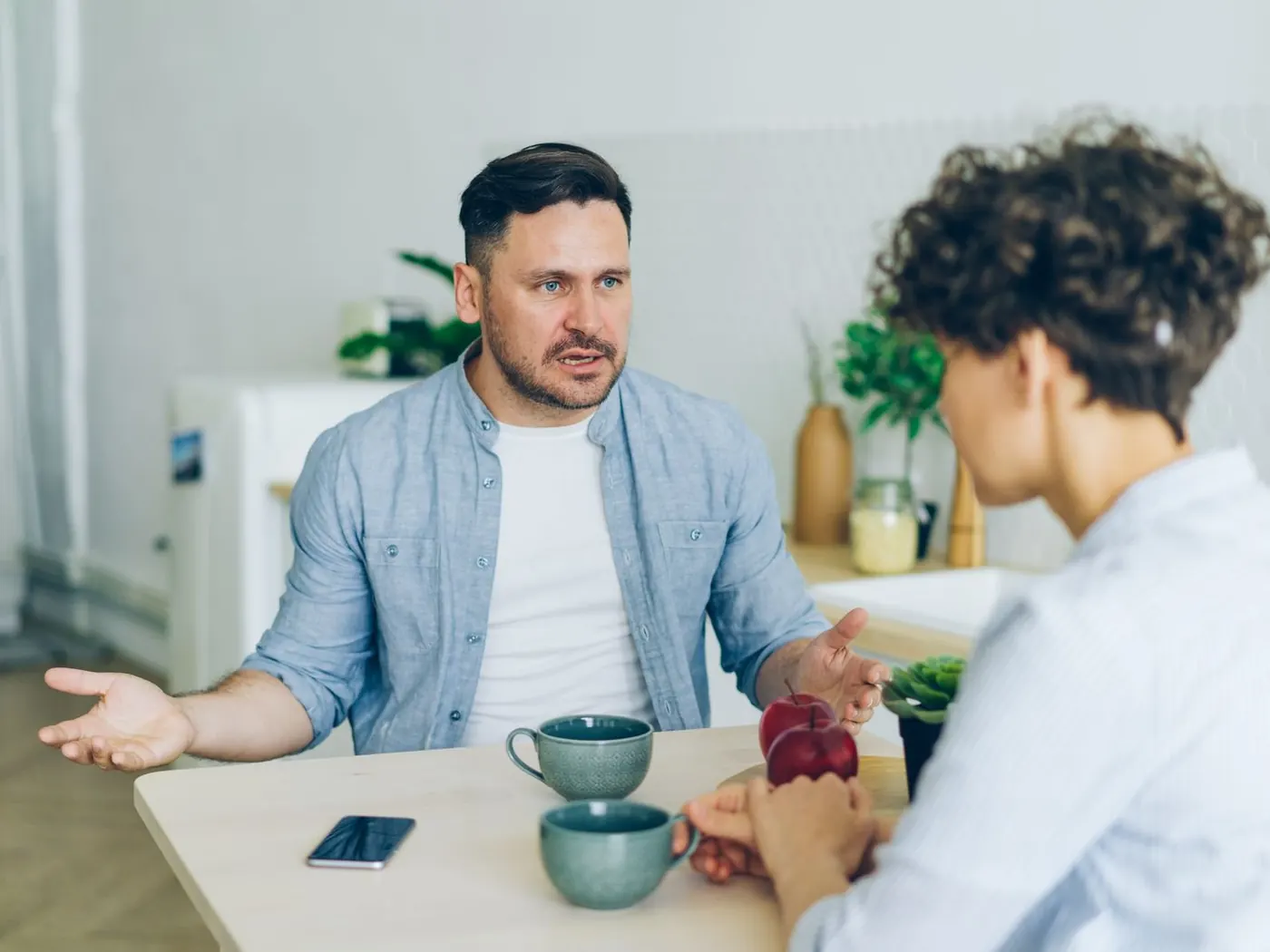 Dos personas conversando sentadas a la mesa de una cocina, con dos tazas entre ellas